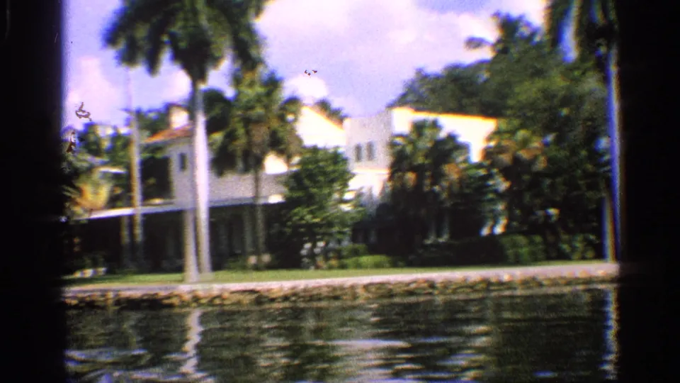 1959 California 8mm frame of a waterside residence, palms, shrubs, lawn, and reflective water.