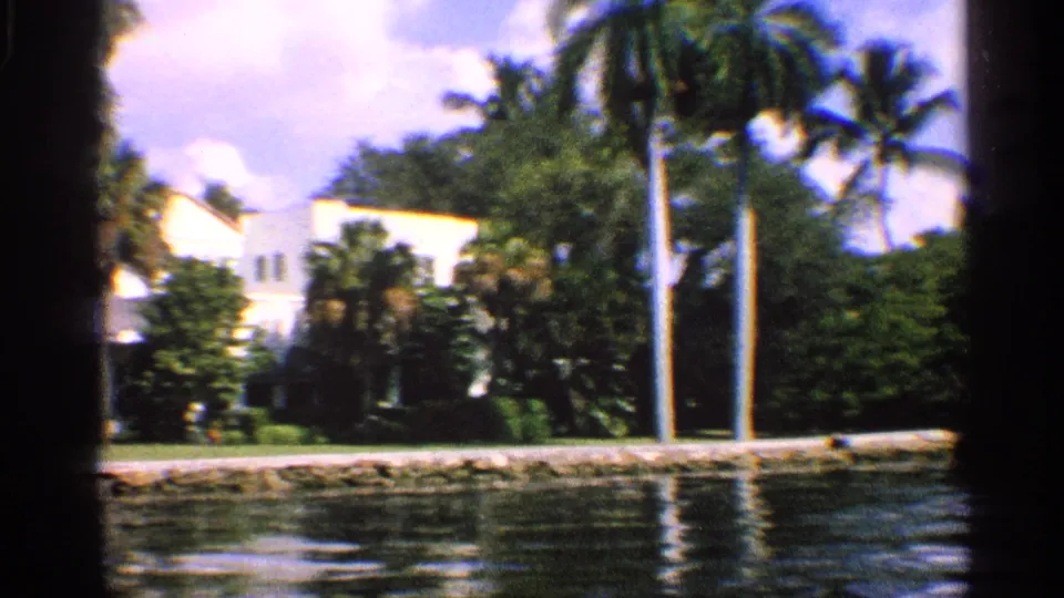 1959 California 8mm frame showing a white house behind palms and foliage along a sunny canal.