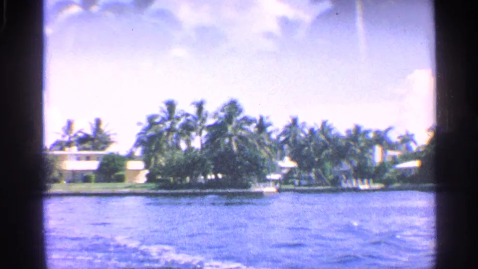 1959 California home-movie frame looking across a canal toward palms and low waterfront houses under bright sky.