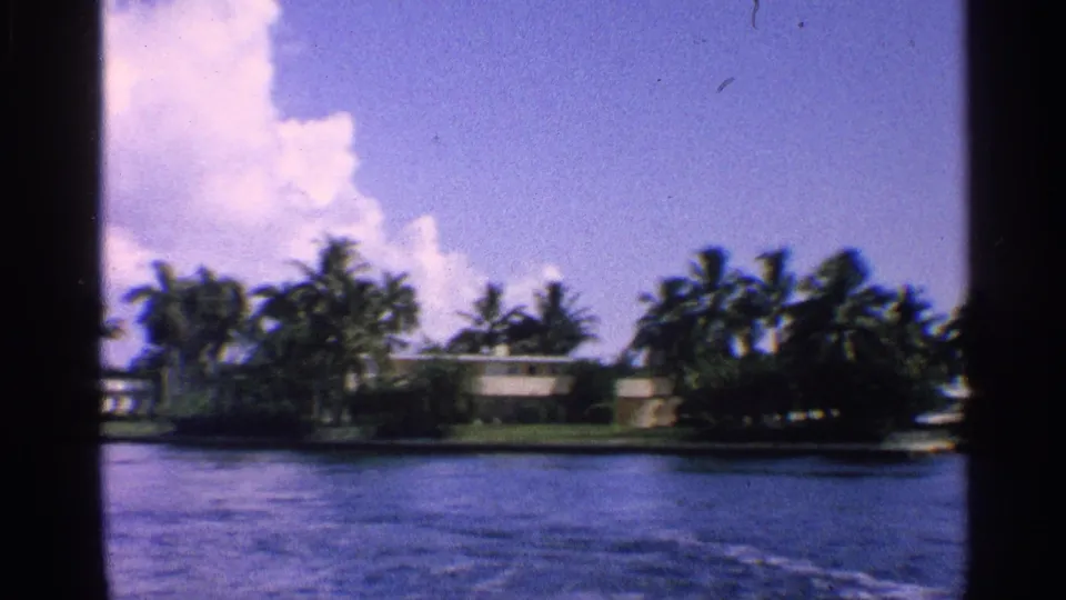 Boat-level 1959 California canal view with a low waterfront house partly framed by palms and a large cloud.