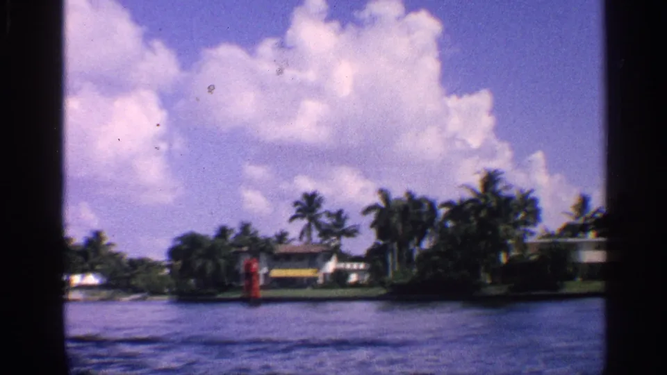 1959 California 8mm frame showing a sunny canal with palms, waterfront homes, clouds, and a red marker near shore.