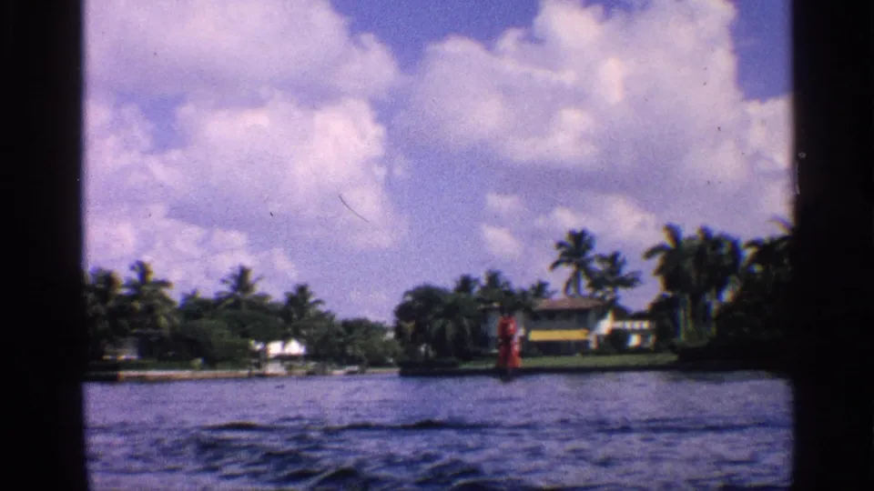 1959 California 8mm frame showing a waterway, palm-lined shore, low houses, bright clouds, and a small red navigation marker.