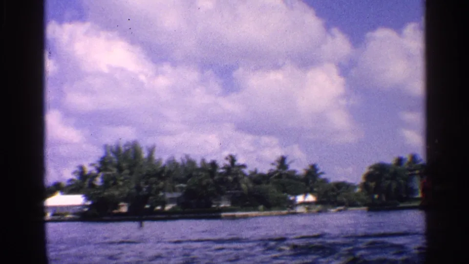 1959 California 8mm frame of a calm waterway passing a tree-lined shore with a white-roofed structure and scattered clouds.