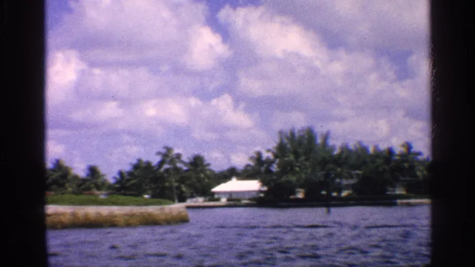 1959 California 8mm frame showing rippled water, a low shoreline, dense trees, a white-roofed building, and bright clouds.