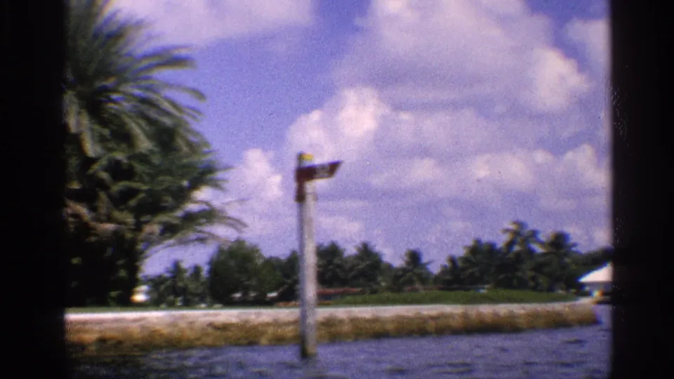 1959 California 8mm frame with a shoreline signpost or marker, palm trees, water, low land, and a bright clouded sky.