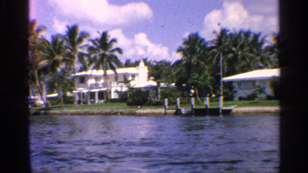 White waterfront home with palm trees, lawn, dock posts, and rippling water.