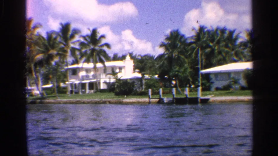 1959 California waterfront scene showing a white mid-century home, palms, manicured lawn, dock posts, and rippling canal water.