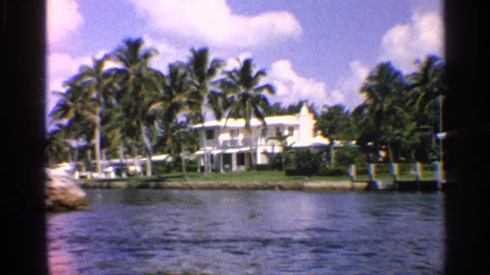 1959 8mm view of a large white waterfront residence in California, framed by palm trees, lawn, shoreline, and blue water.