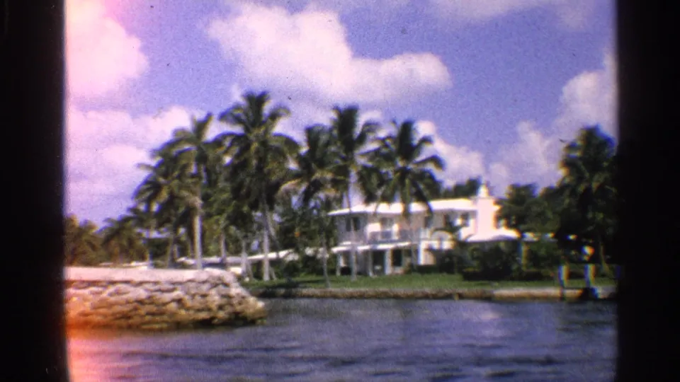 1959 California waterway scene with a rocky shoreline in front, palm-lined homes across the water, and bright clouds overhead.