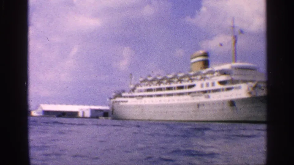 The liner is seen lower and closer in frame, with blue water and a pale dock structure at the left.