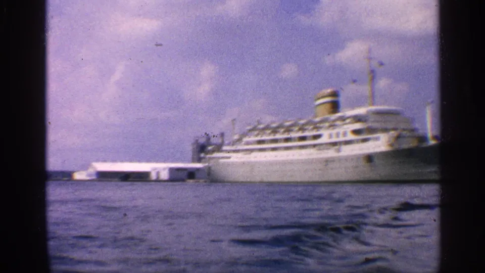 1959 California 8mm still of a docked white passenger ship, low shoreline structure, and rippled harbor water in the foreground.