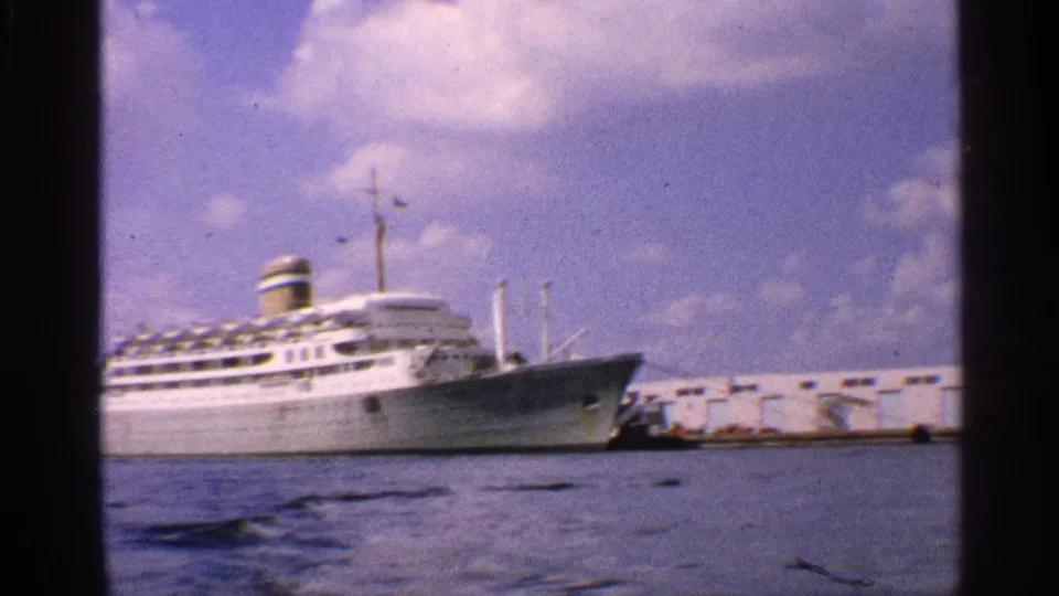 The camera continues across the 1959 California port, holding on the docked white passenger ship and pier.