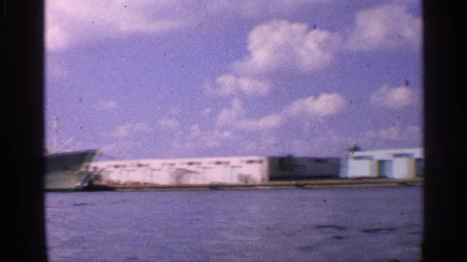 A California port waterfront appears with long low buildings, open water, and part of the docked ship at the left edge.