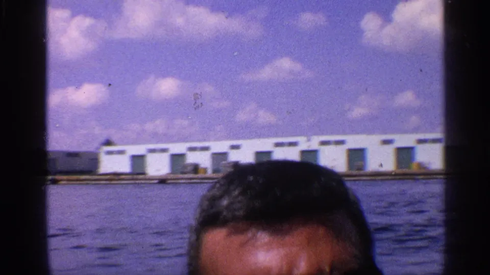 An adult man wearing dark sunglasses appears in the foreground at a 1959 California port, with water and low buildings behind him.