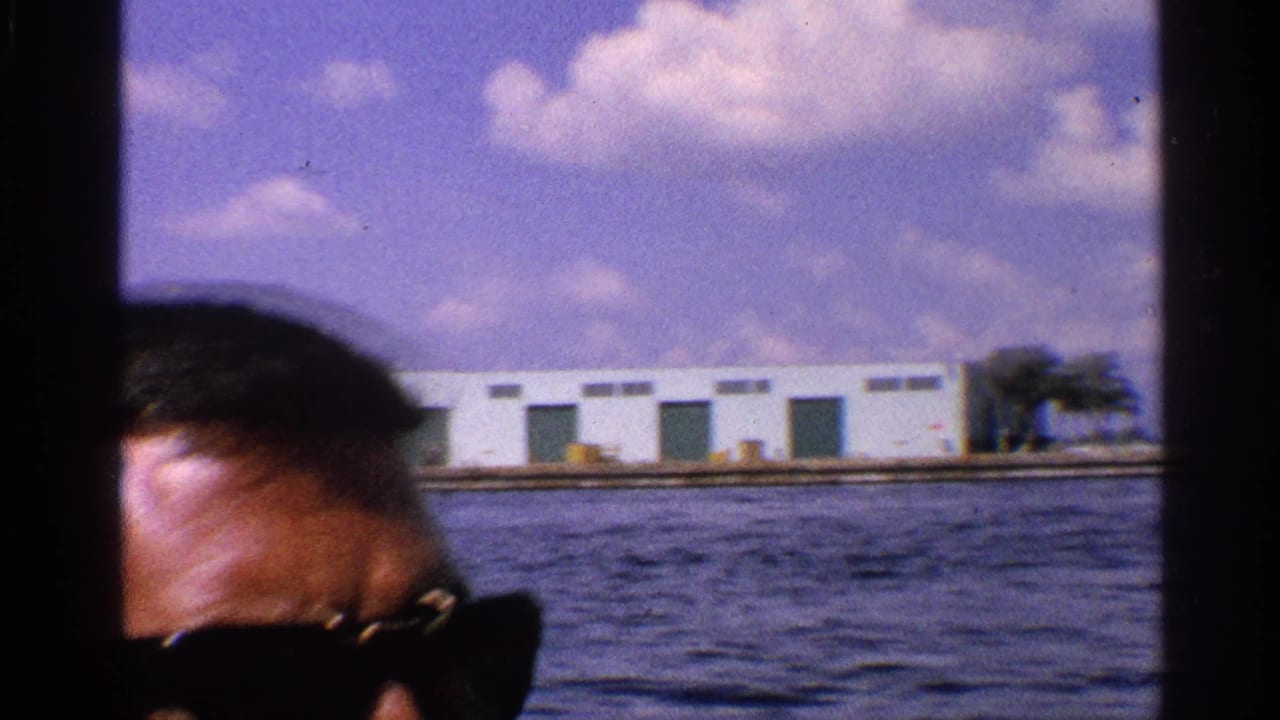 Partial close view of a man in dark sunglasses during a 1959 California boat ride, with water, low buildings, and clouds beyond him.