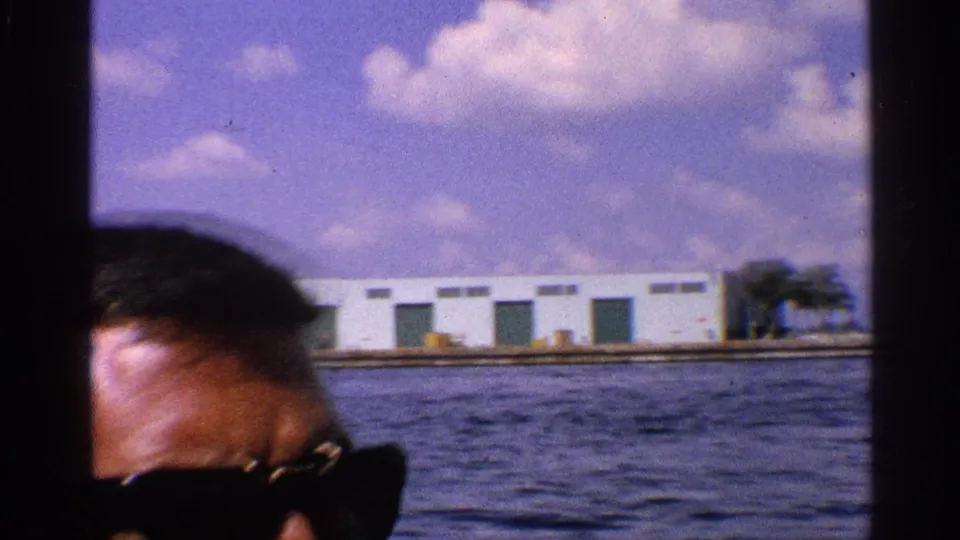 Partial close view of a man in dark sunglasses during a 1959 California boat ride, with water, low buildings, and clouds beyond him.