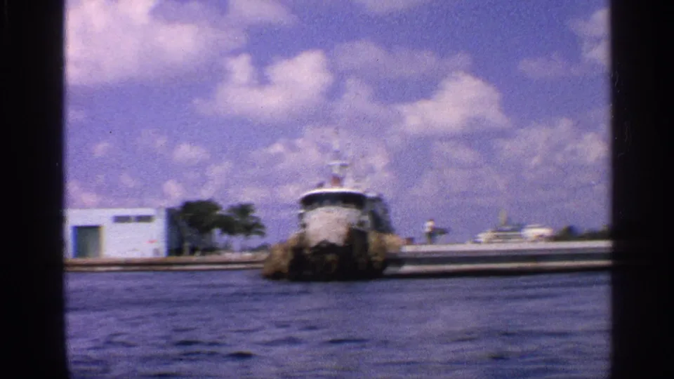 A tugboat moves across the water near low white waterfront buildings in the supplied 1959 California home-movie footage.