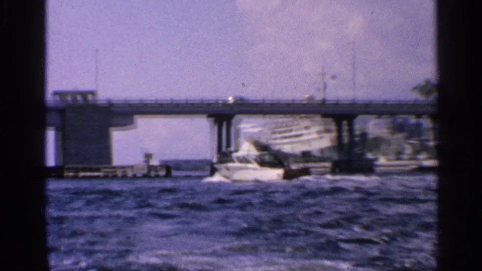 A large white ship appears near or beneath a low bridge, filmed from across the water in the 1959 California home-movie clip.