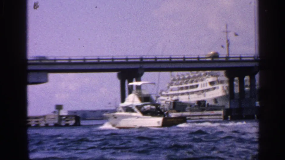 1959 California home-movie frame showing a white motorboat passing near a bridge while a larger passenger vessel sits in the background.