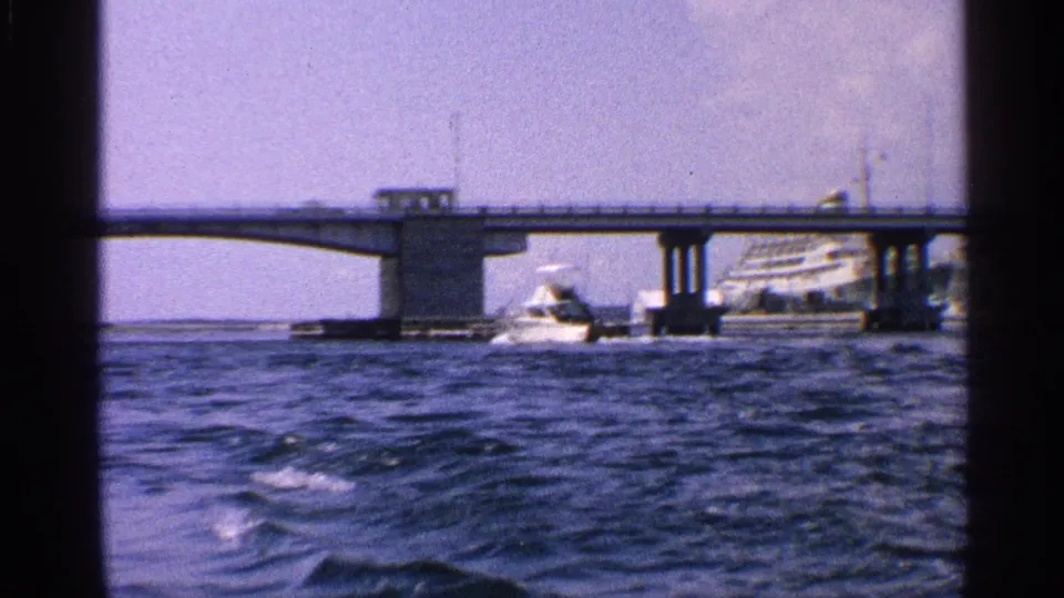 Wide 1959 waterfront view with bridge spans, a white motorboat, choppy water, and a large passenger vessel behind the bridge.
