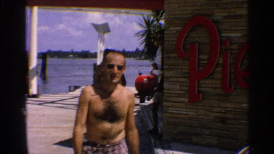Man in sunglasses and swim trunks stands on a dock near waterfront signage in a 1959 California home-movie frame.