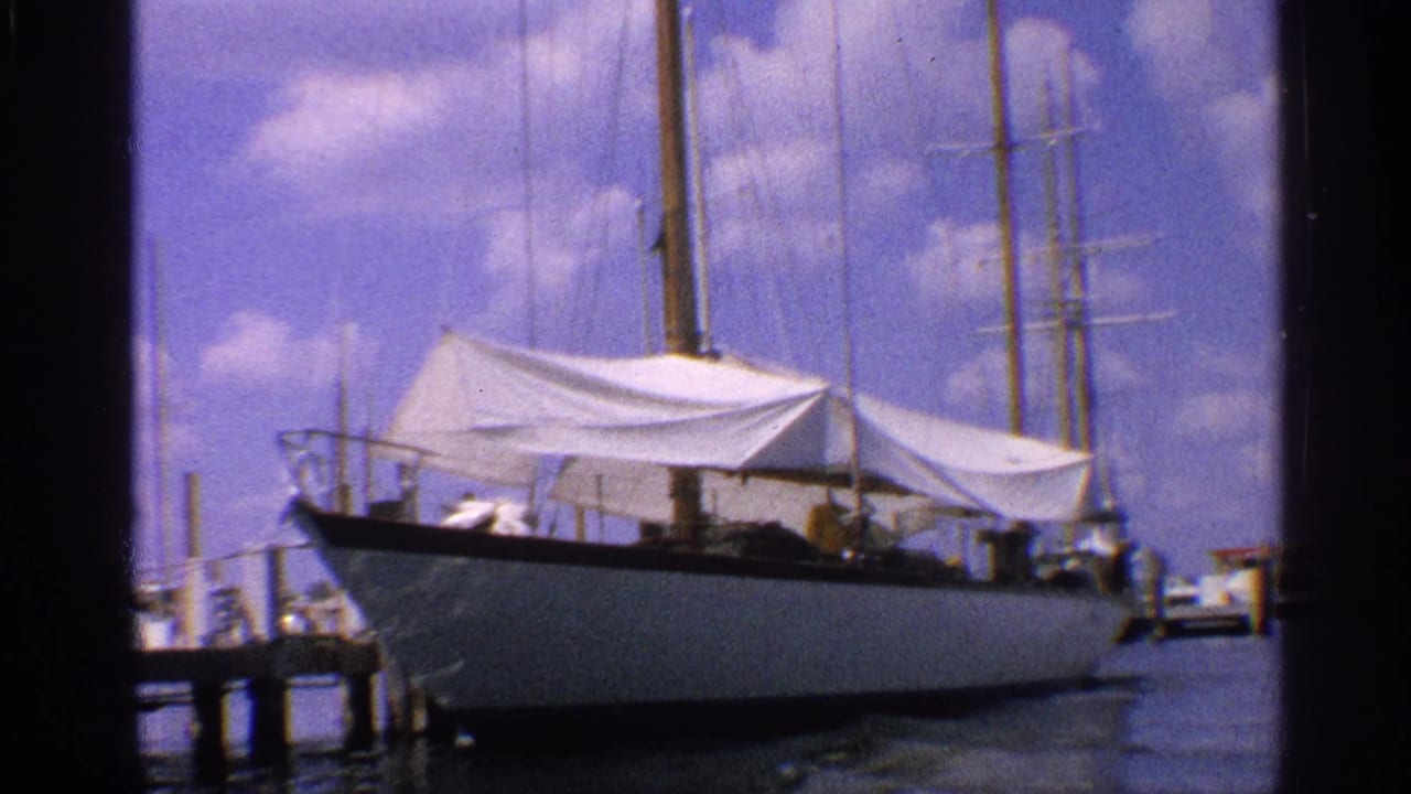 1959 California home-movie frame of a tarp-covered sailboat at a pier, with tall masts rising into a clouded blue sky.
