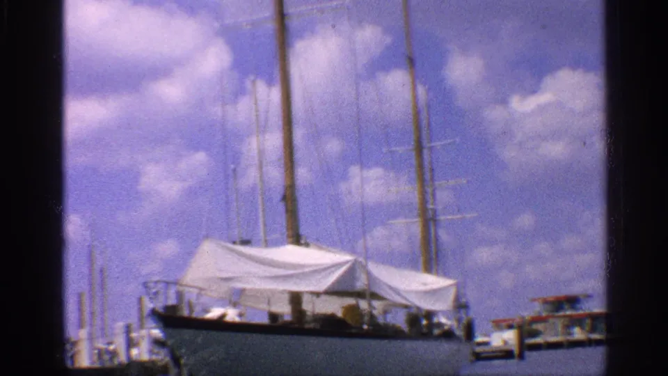 The covered sailboat remains docked at the pier, with rigging, mast lines, blue sky, and possible distant figures near the vessel.