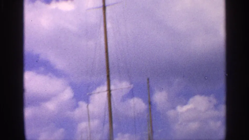 A handheld upward view emphasizes the sailboat mast and rigging against a soft blue sky with scattered clouds.