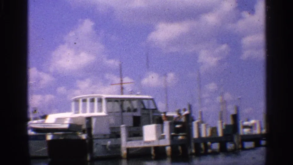 White cabin boat moored at a California marina pier, with masts and dock posts visible in 1959 home-movie footage.