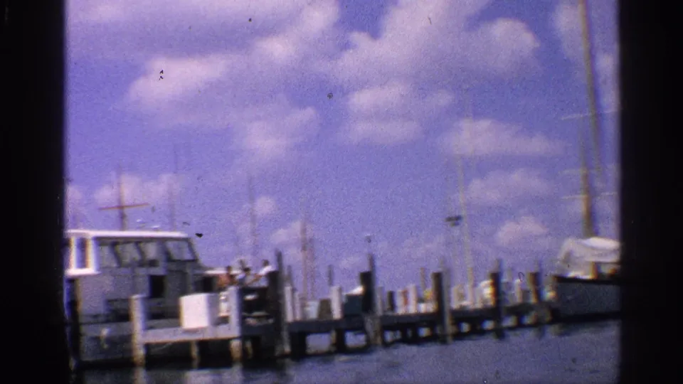 California marina view with a white boat, wooden pier, masts, water, and distant people in 1959 8mm footage.