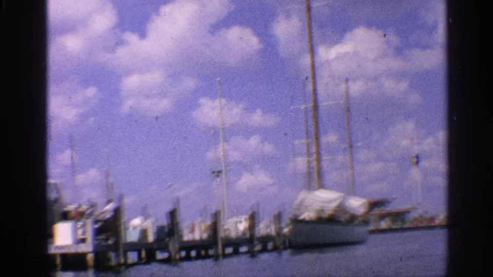 Wide marina frame in 1959 California home-movie footage, showing masts, dock posts, water, and a covered moored boat.