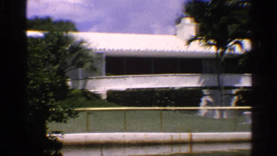 1959 California 8mm frame showing a white modernist building partly obscured by trees, hedges, and a waterfront barrier.