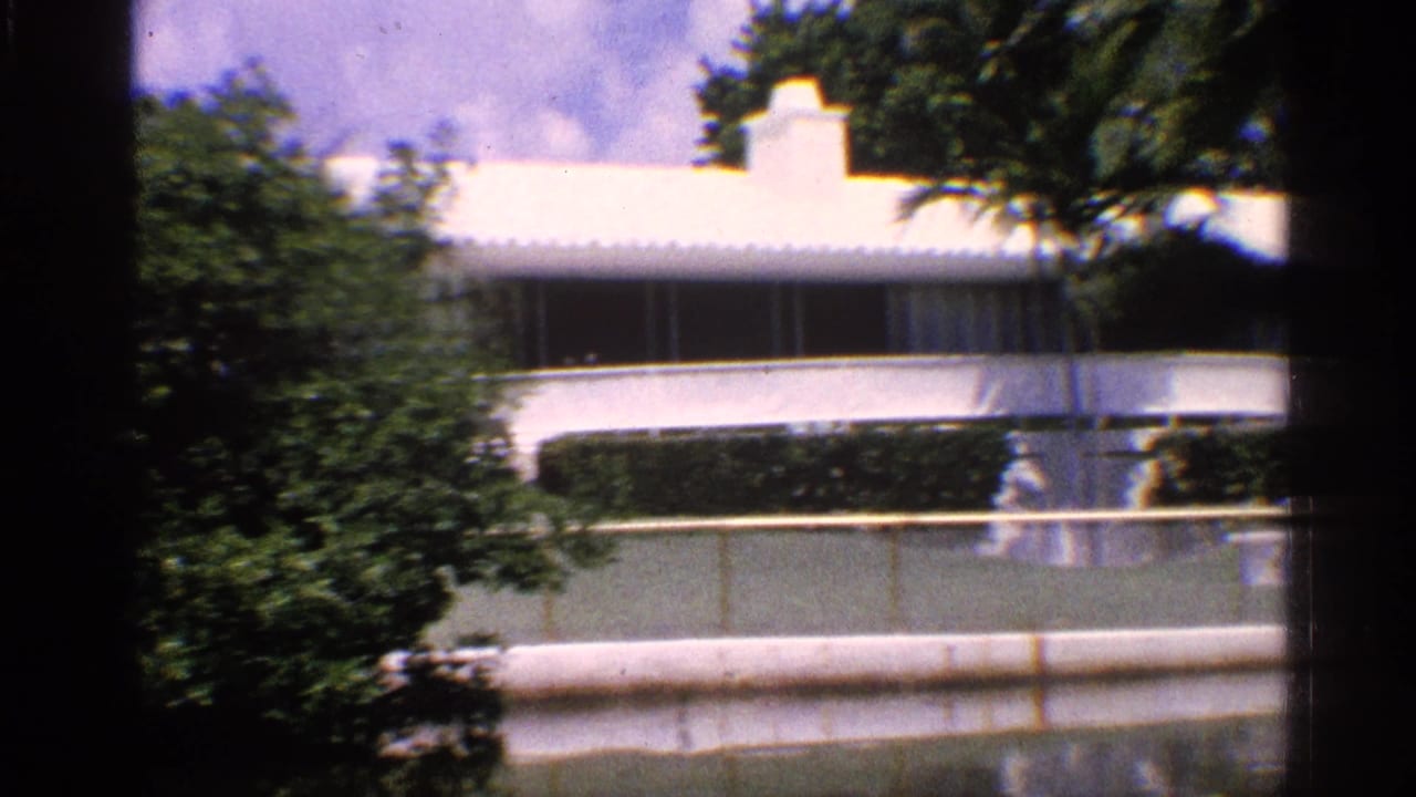 White building, trees, hedges, and railings seen from across a barrier.
