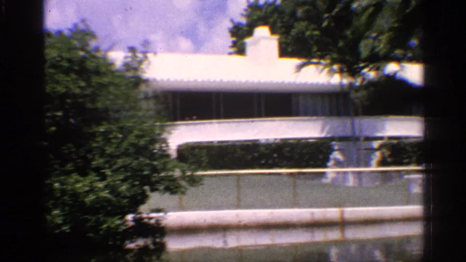 1959 California 8mm frame with a white modernist building behind greenery and a pale waterfront railing under a blue sky.
