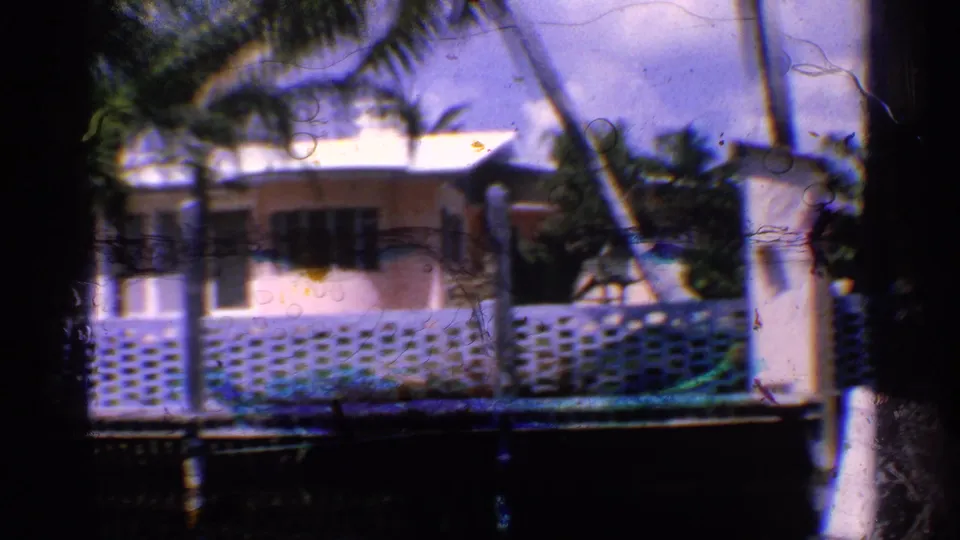 1959 California 8mm frame showing a pink bungalow, palm trees, a patterned blue wall, and waterfront posts in bright daylight.