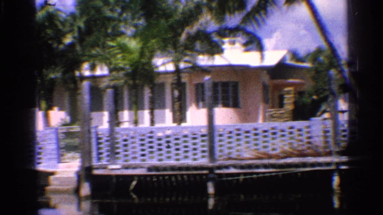 1959 California 8mm frame showing a light waterfront house, palm trees, white block fencing, and a dock or seawall beside calm water.