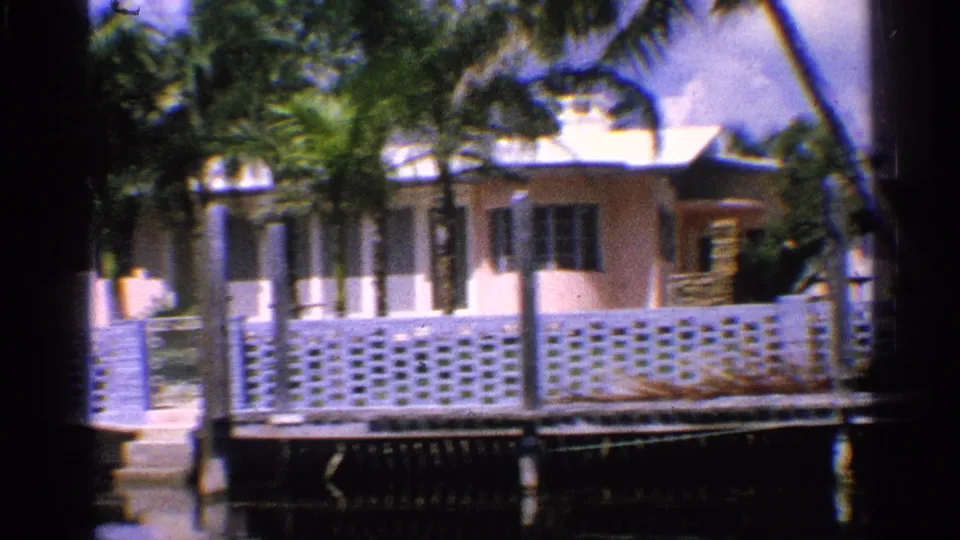 1959 California 8mm frame showing a light waterfront house, palm trees, white block fencing, and a dock or seawall beside calm water.