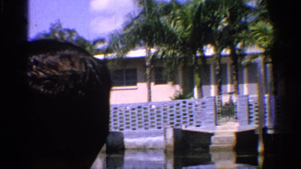 1959 California 8mm frame with a partial person's head in foreground and a palm-lined waterfront house behind white fencing.