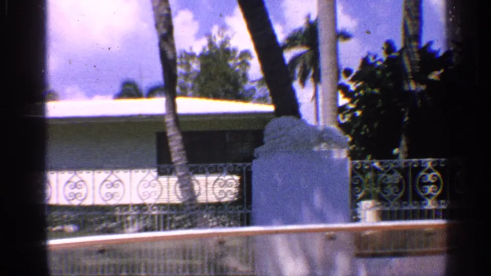 1959 California 8mm frame showing a low waterfront house, decorative metal railing, white wall, palm trunks, and bright sky.