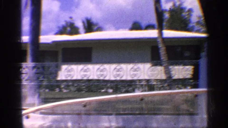 1959 California 8mm frame of a waterfront residence with palm trunks, patterned wall panels, roofline, and water at the lower edge.