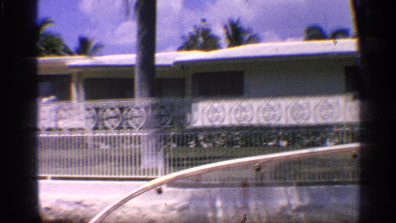 1959 California home-movie frame showing a low ranch-style house behind ornate fencing, seen past a vehicle window and canal edge.