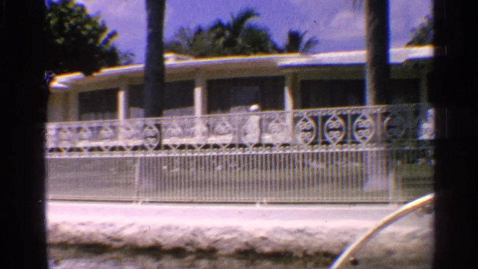 1959 California suburban exterior with a white decorative fence, ranch-style house, palm trunks, and a concrete canal edge.