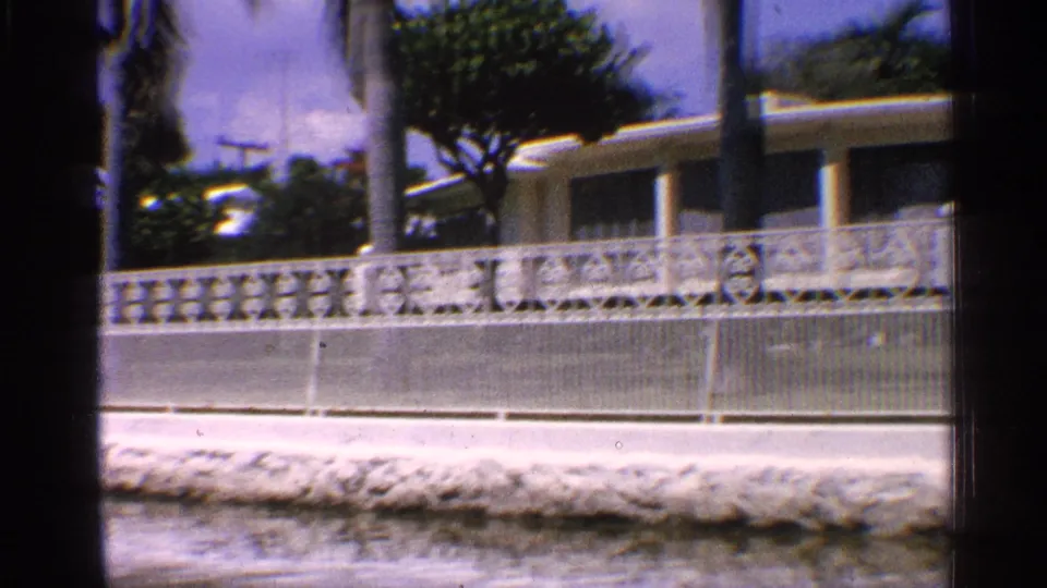 1959 California frame of a canal-side ranch-style residence with ornate fencing, palms, water, and dark 8mm gate edges.