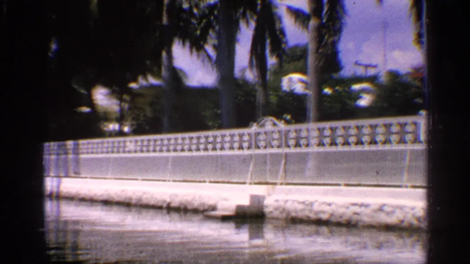 1959 California home-movie view of decorative canal-side fencing, palms, greenery, and a partly obscured residential exterior.
