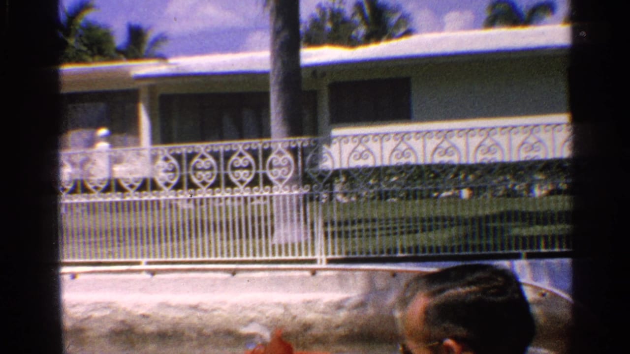 1959 California 8mm frame of a passenger partly seen below a white house, decorative railings, palm trees, and a water feature.