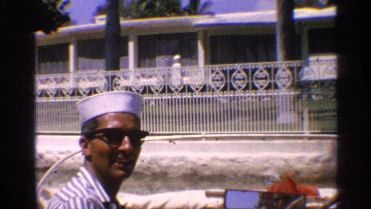 Man in a white sailor-style hat and sunglasses rides past a white house and palm trees.