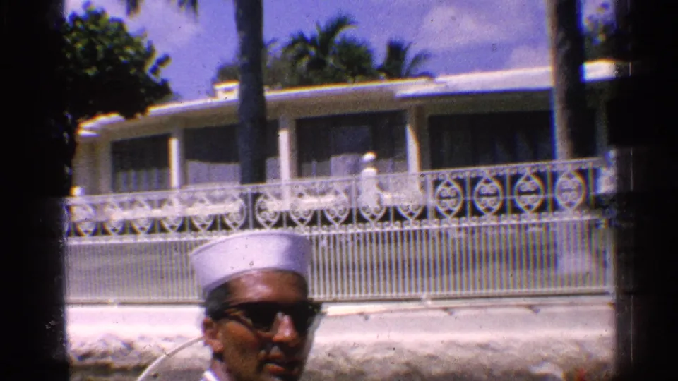 1959 California home-movie frame with a man in sunglasses and white cap below a sunlit house, railings, palms, and a pale wall.