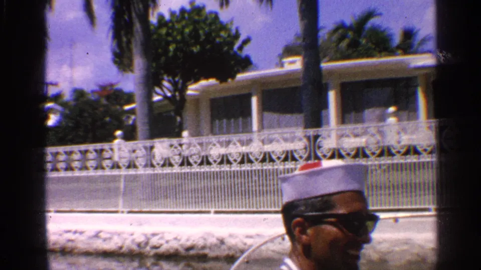 1959 California 8mm frame showing the man in profile wearing a white cap and sunglasses as palms and a white house pass behind him.
