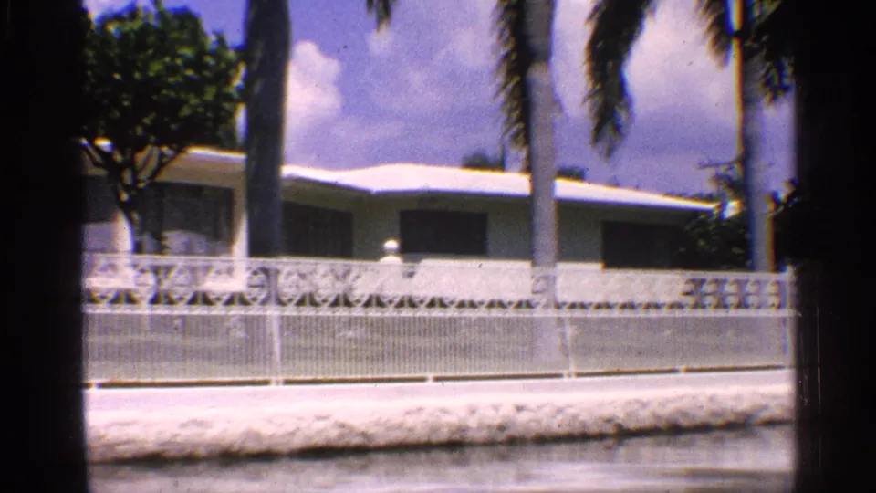 Vintage 8mm view of a California residential exterior with a white flat-roof house, shaded windows, palms, and a decorative fence.