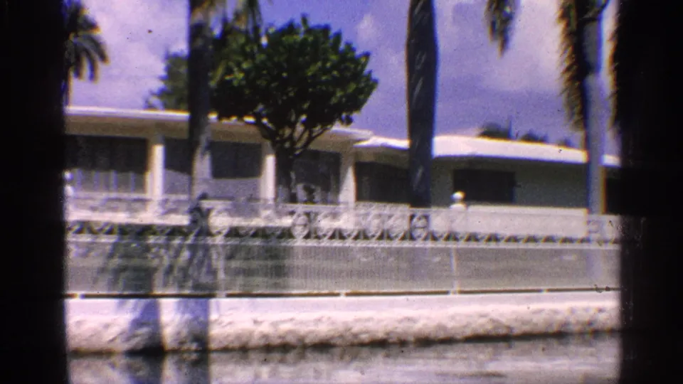 California 1959 home-movie frame showing a white low house, palm trees, decorative fence, and dark 8mm frame edges.